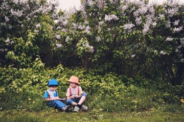 Fotografia raffigurante due bambini seduti in un prato, vestiti con abiti da giardino